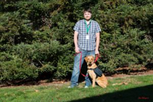 Photo of Josh and Mim who is wearing a blue service dog vest stand in front of redwood branches